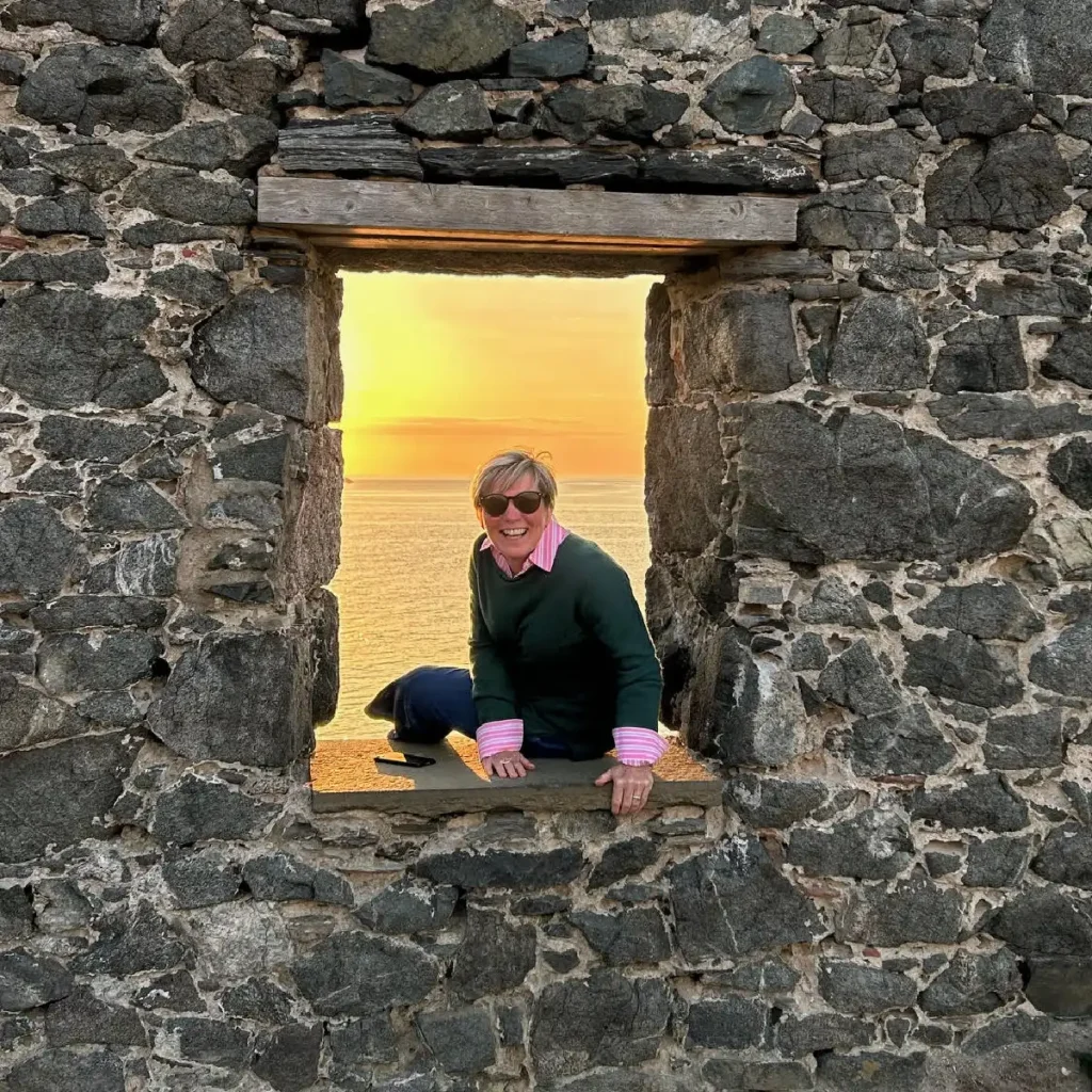 A smiling woman wearing sunglasses (Donna Ronald) sits on the window sill of a ruined building, the setting sun visible in the open window behind her.