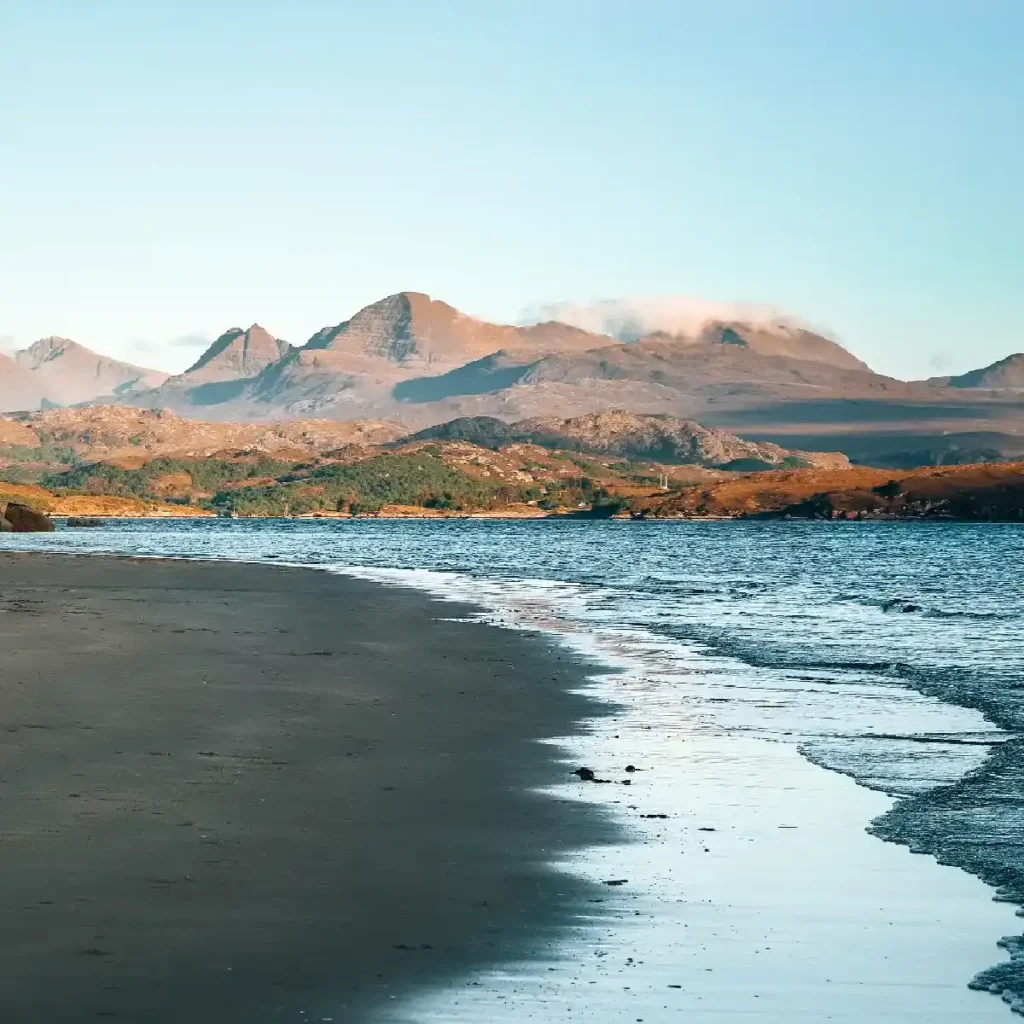 A beach of dark sand with tall mountains behind on Scotland's West Coast. Image by Torin Liversedge
